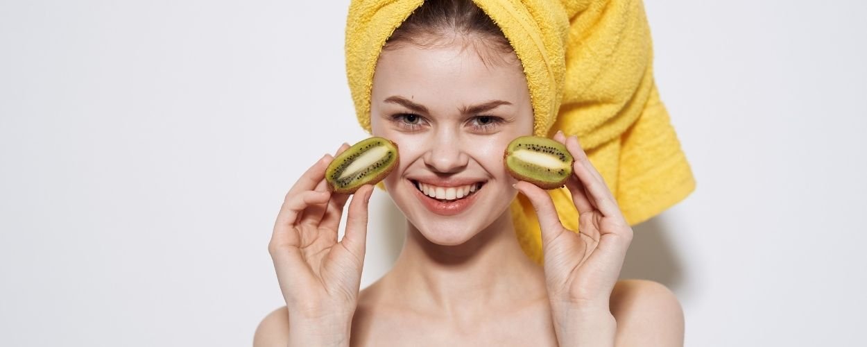 Woman with a towel on her head and two half kiwis in her hands that she holds against her cheeks